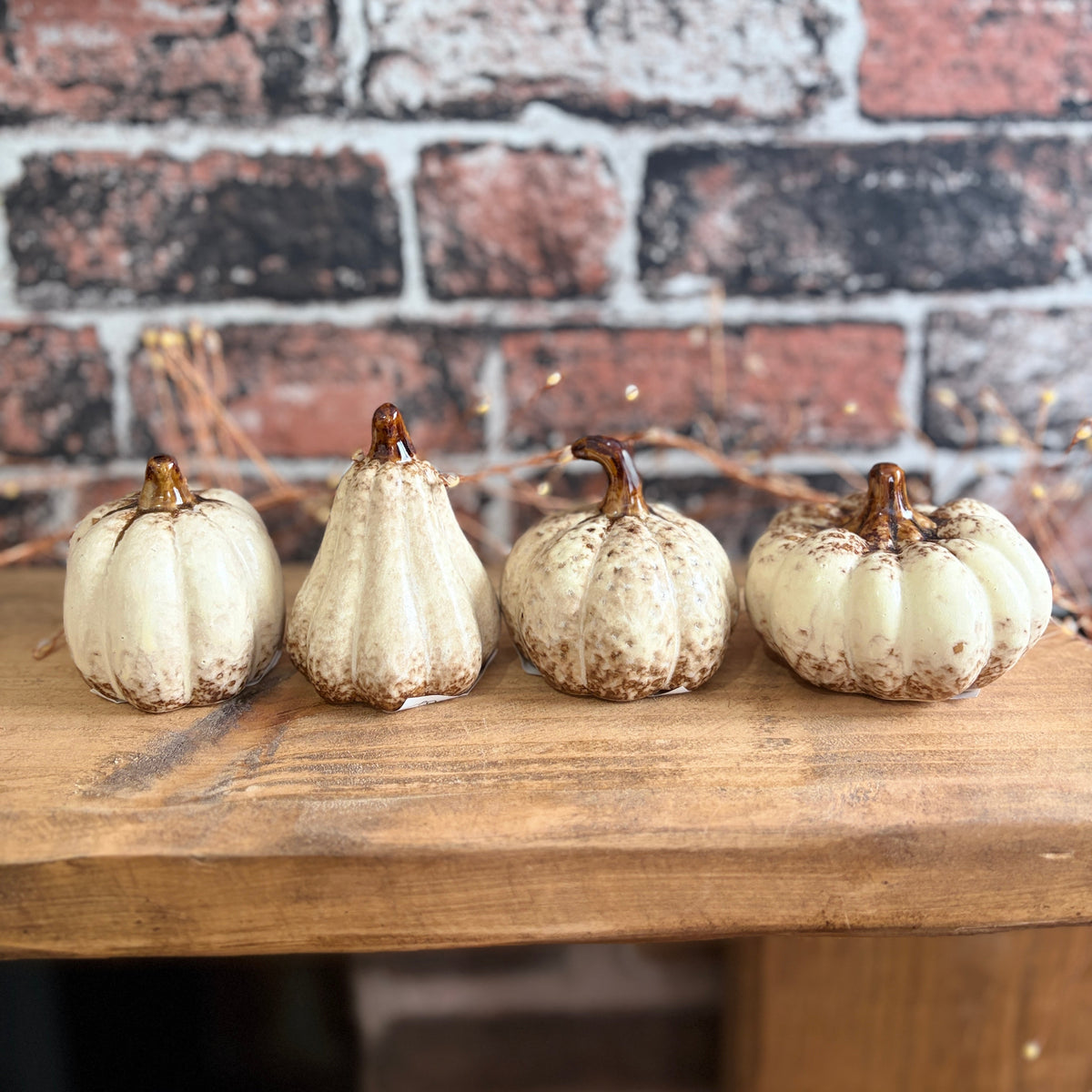 Four small decorative pumpkins in off-white and beige tones with brown stems, lined up on a wooden surface against a rustic red and gray brick wall background.