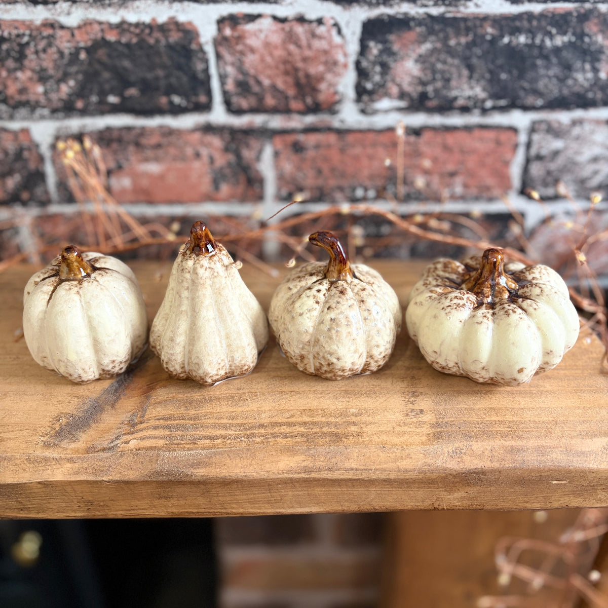 Four small decorative pumpkins in off-white and cream tones with brown stems, arranged in a row on a wooden shelf against a rustic red and black brick wall.