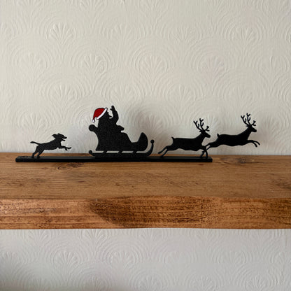 A black recycled plastic Christmas silhouette decoration of Santa Claus in a sleigh pulled by two reindeer and a dog, displayed on a rustic wooden shelf against a textured off-white wall.