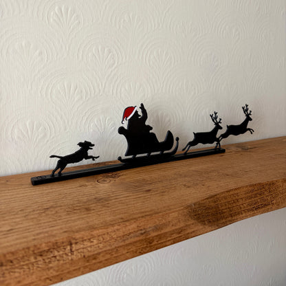 A black recycled plastic Christmas shelf decor piece featuring Santa Claus in a sleigh pulled by three reindeer and a dog, displayed on a wooden mantel against a textured off-white wall.