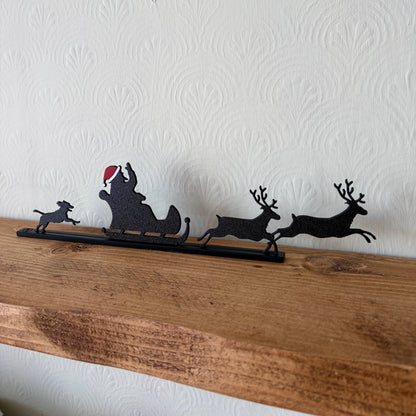 A black recycled plastic Christmas silhouette decoration featuring Santa Claus in a sleigh with reindeer and a dog, displayed on a rustic wooden mantel against a white textured wall.
