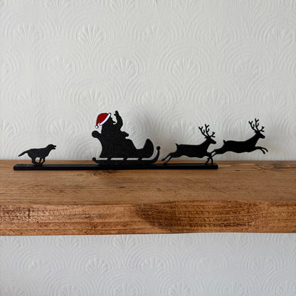 A black recycled plastic Christmas decor piece featuring Santa Claus in a sleigh pulled by two reindeer, with a small black dog figure on the left, displayed on a rustic wooden shelf against a white textured wall.