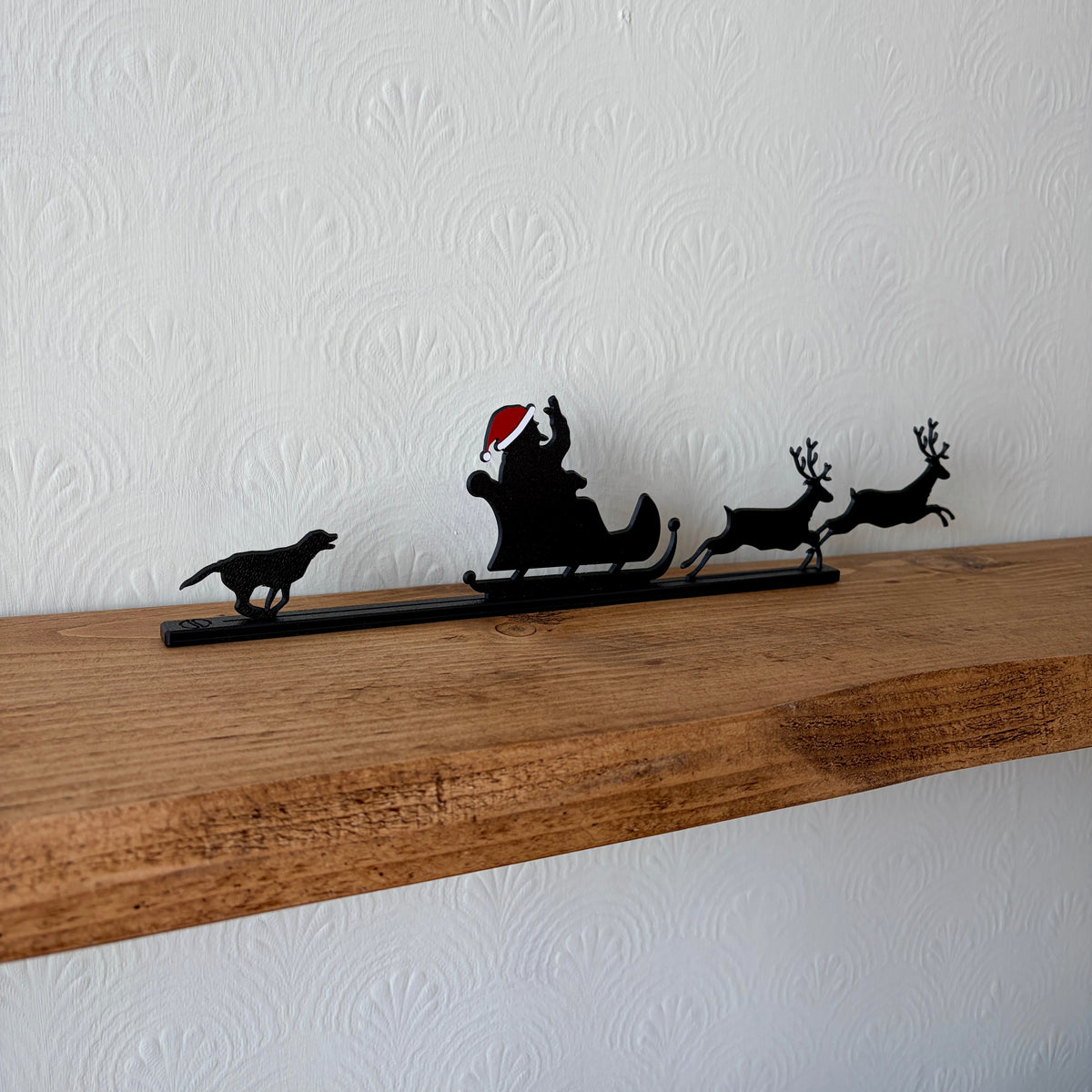 A black recycled plastic Christmas shelf decor piece featuring a silhouette of Santa Claus in a sleigh pulled by two reindeer, with a small dog figure on the left, displayed on a rustic wooden shelf against a white textured wall.