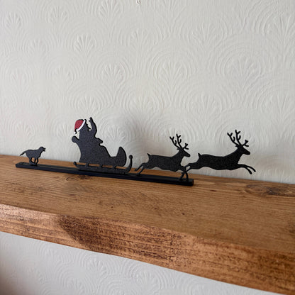 A black recycled plastic silhouette Christmas decor piece featuring Santa Claus in a sleigh pulled by two reindeer, with a small dog on the left, displayed on a rustic wooden shelf against a white textured wall.