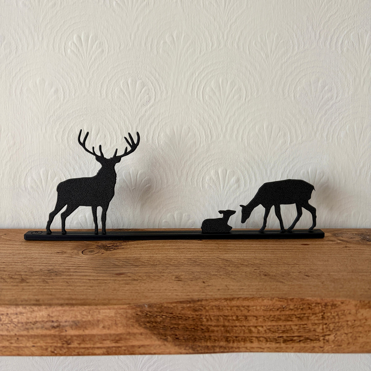 Black recycled plastic deer silhouette shelf decor featuring a stag, a doe, and a fawn on a long horizontal bar, displayed on a rustic wooden mantel against a textured off-white wall.