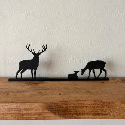 Black recycled plastic deer silhouette shelf decor featuring a stag, a doe, and a fawn on a long horizontal bar, displayed on a rustic wooden mantel against a textured off-white wall.