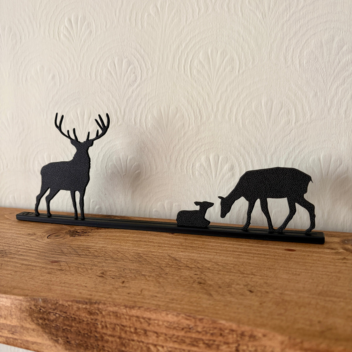 Black recycled plastic deer silhouette shelf decor featuring a stag, a doe with fawn, and a long horizontal base, displayed on a wooden mantel against a textured off-white wall.