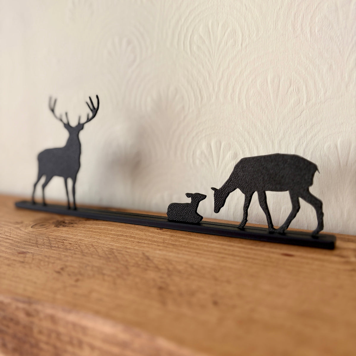A black recycled plastic deer silhouette shelf decor piece featuring a stag, a doe with fawn, and a long base, displayed on a wooden surface against a textured cream wall.