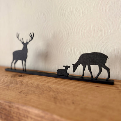 A black recycled plastic deer silhouette shelf decor piece featuring a stag, a doe with fawn, and a long base, displayed on a wooden surface against a textured cream wall.