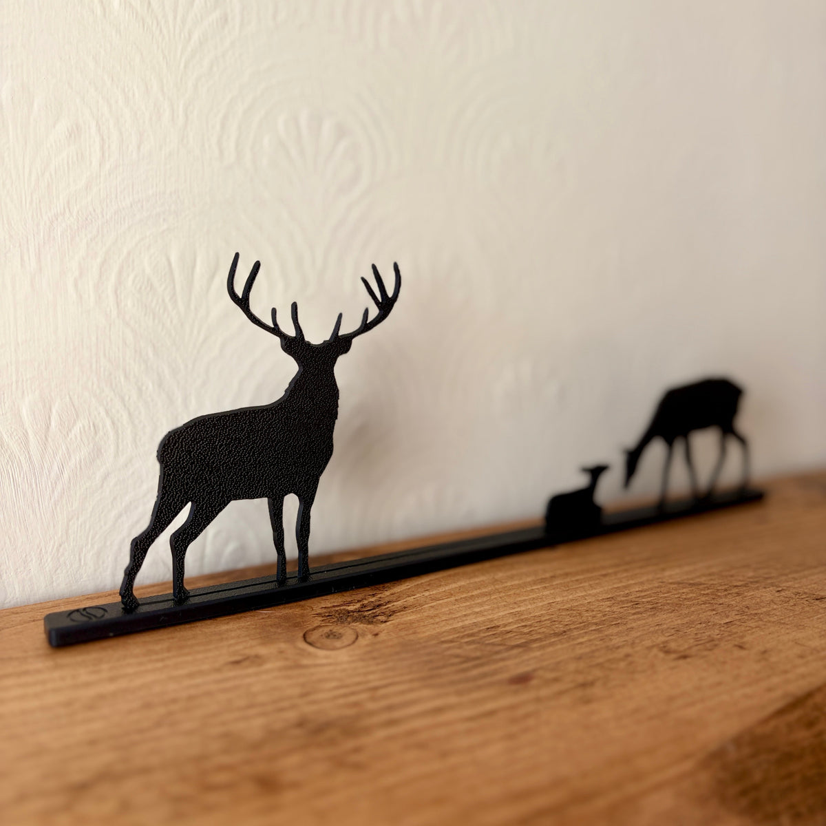 A black recycled plastic deer silhouette shelf mount with a stag, doe, and fawn design, displayed on a wooden shelf against a textured off-white wall.