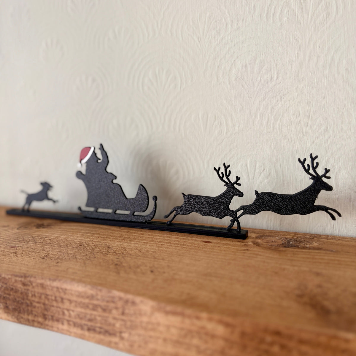 A black recycled plastic Christmas shelf decoration featuring Santa Claus in a sleigh with reindeer, displayed on a rustic wooden mantel against a textured off-white wall.