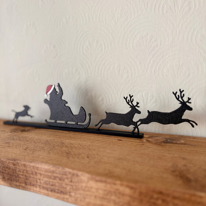 A black recycled plastic Christmas shelf decoration featuring Santa Claus in a sleigh with reindeer, displayed on a rustic wooden mantel against a textured off-white wall.