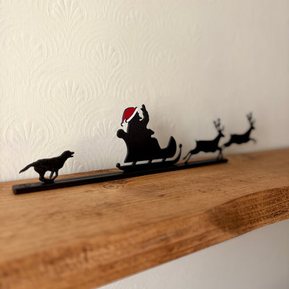A black recycled plastic Christmas shelf silhouette featuring Santa Claus in a sleigh pulled by two reindeer, with a dog on the left, displayed on a rustic wooden shelf against a textured off-white wall.