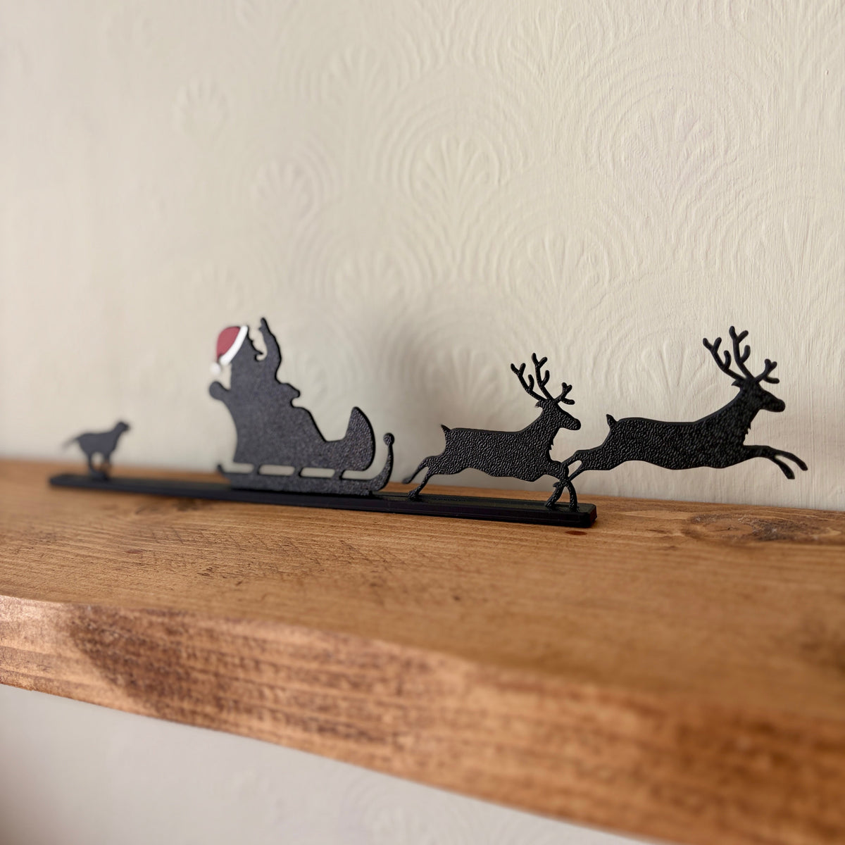 A black recycled plastic Christmas shelf silhouette featuring Santa Claus in a sleigh with reindeer, displayed on a rustic wooden mantel against a textured off-white wall.