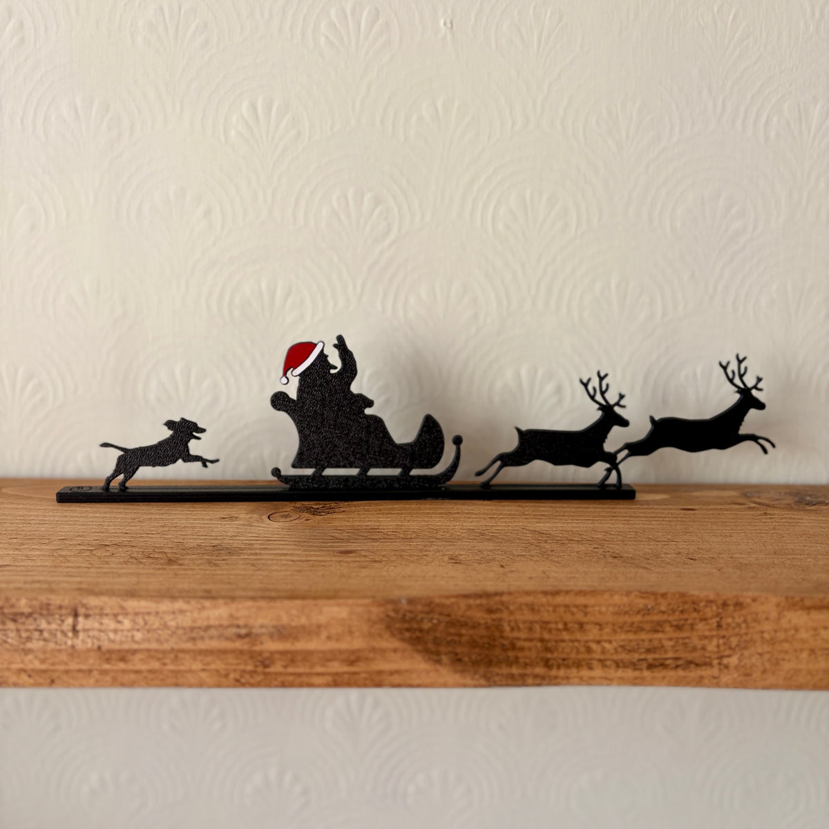 A black recycled plastic silhouette holiday shelf decor featuring Santa Claus in a sleigh with reindeer and a dog, wearing a red Santa hat, displayed on a rustic wooden mantel against a textured off-white wall.