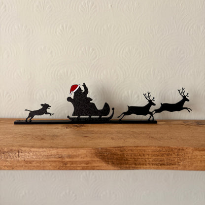 A black recycled plastic silhouette holiday shelf decor featuring Santa Claus in a sleigh with reindeer and a dog, wearing a red Santa hat, displayed on a rustic wooden mantel against a textured off-white wall.