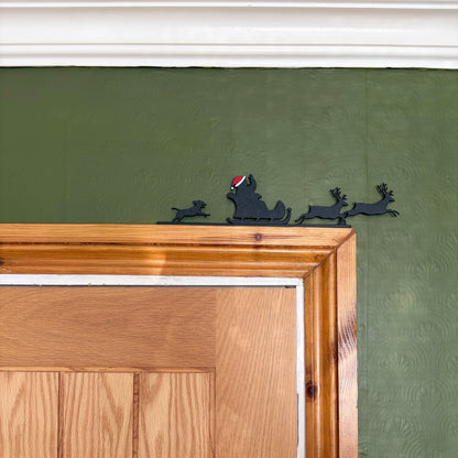 A black recycled plastic silhouette wall decor piece of Santa Claus and his sleigh with reindeer, mounted on a textured olive green wall above a wooden door frame.