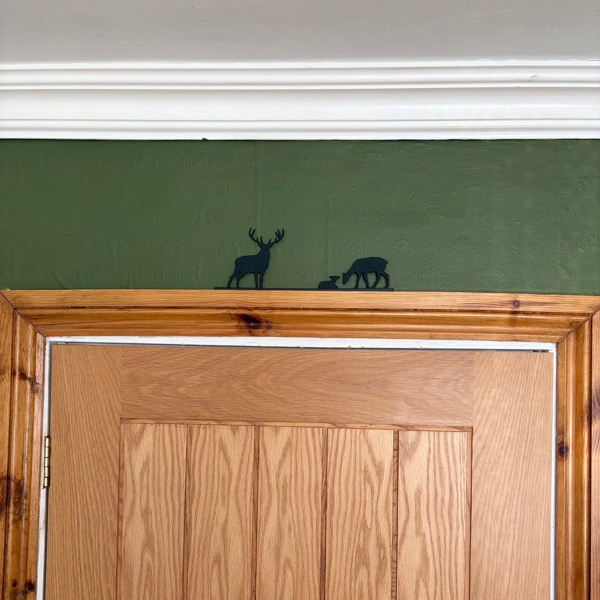 A black deer silhouette door topper of a stag and a fawn mounted on a green wall above a wooden door.