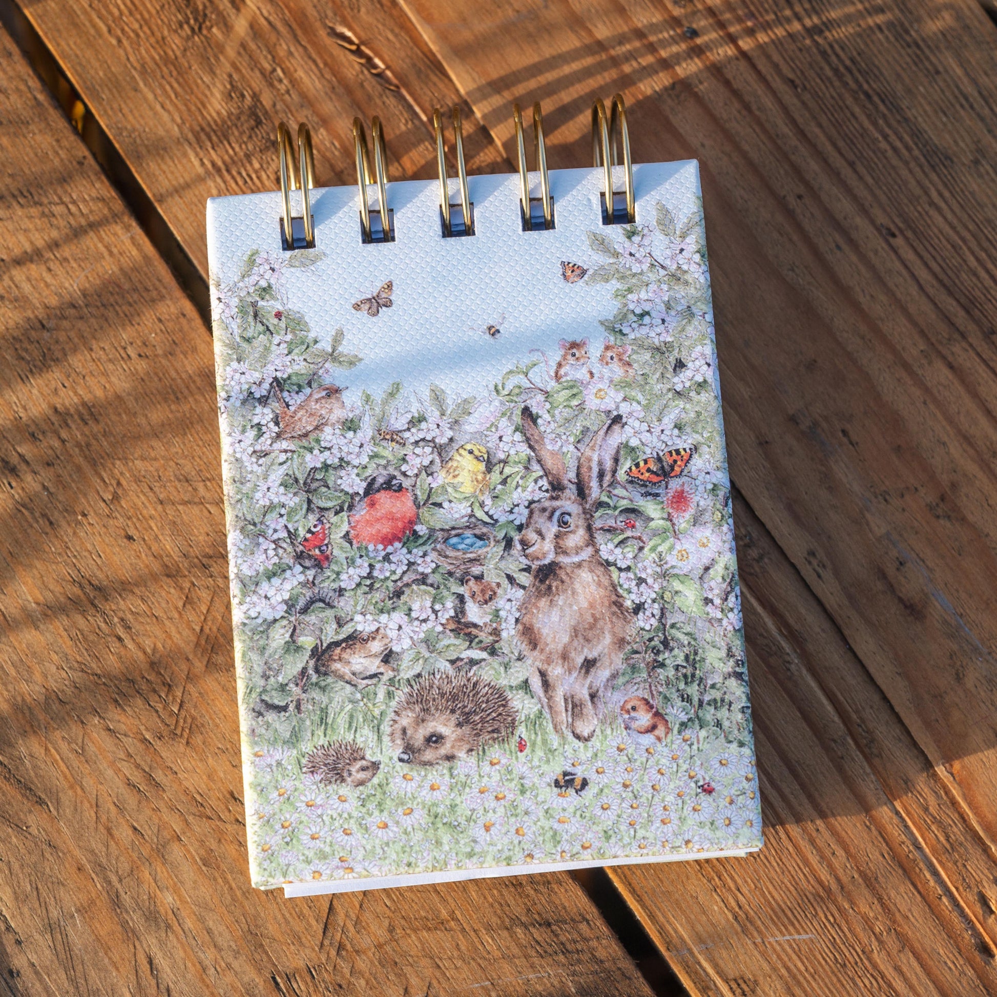 A small spiral-bound notebook with a colorful woodland cover illustration of a hare, hedgehog, flowers, and birds, placed on a rustic wooden surface.