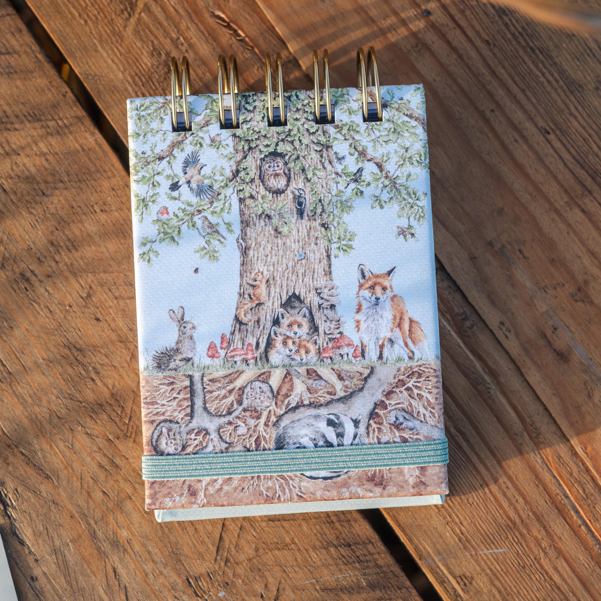 A small spiral-bound notebook with a woodland forest cover illustration of a tree and foxes, placed on a rustic wooden surface.