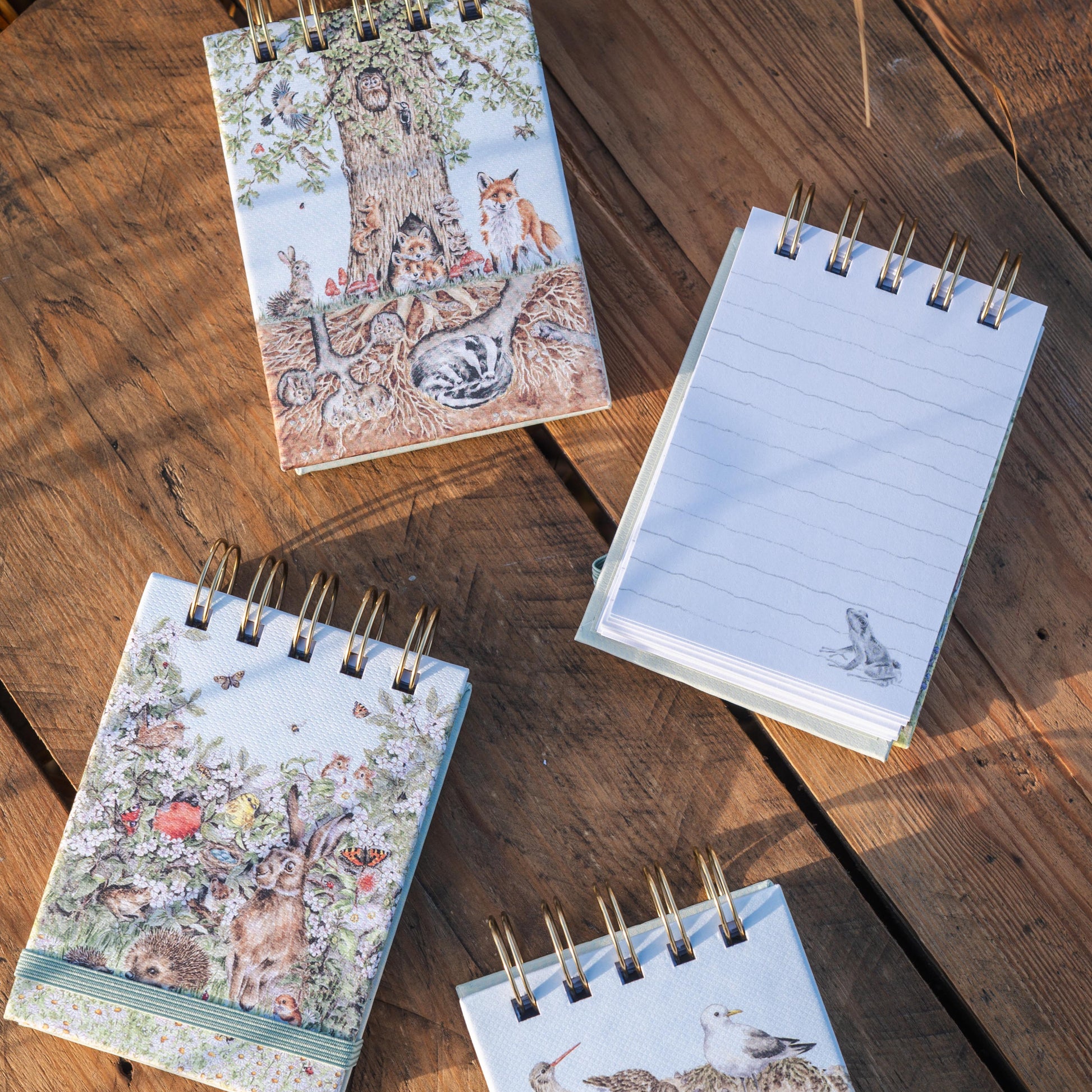 Four small spiral-bound notebooks with woodland illustrations are arranged on a rustic wooden surface. One notebook is open, showing lined pages, while the others are closed with covers featuring trees, birds, and floral designs.