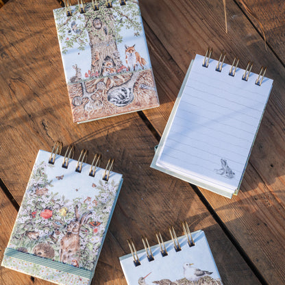 Four small spiral-bound notebooks with woodland illustrations are arranged on a rustic wooden surface. One notebook is open, showing lined pages, while the others are closed with covers featuring trees, birds, and floral designs.