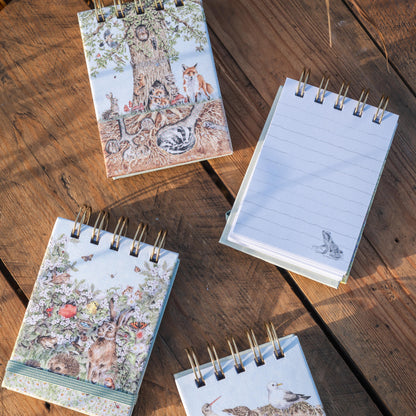 Four small spiral-bound notebooks with woodland illustrations are arranged on a rustic wooden surface. One notebook is open, showing lined pages, while the others are closed with covers featuring trees, birds, and floral designs.
