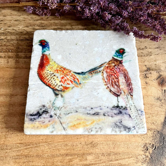 A square natural marble coaster with 2 watercolour painted pheasants displayed on a shelf with purple dried flowers as decoration.