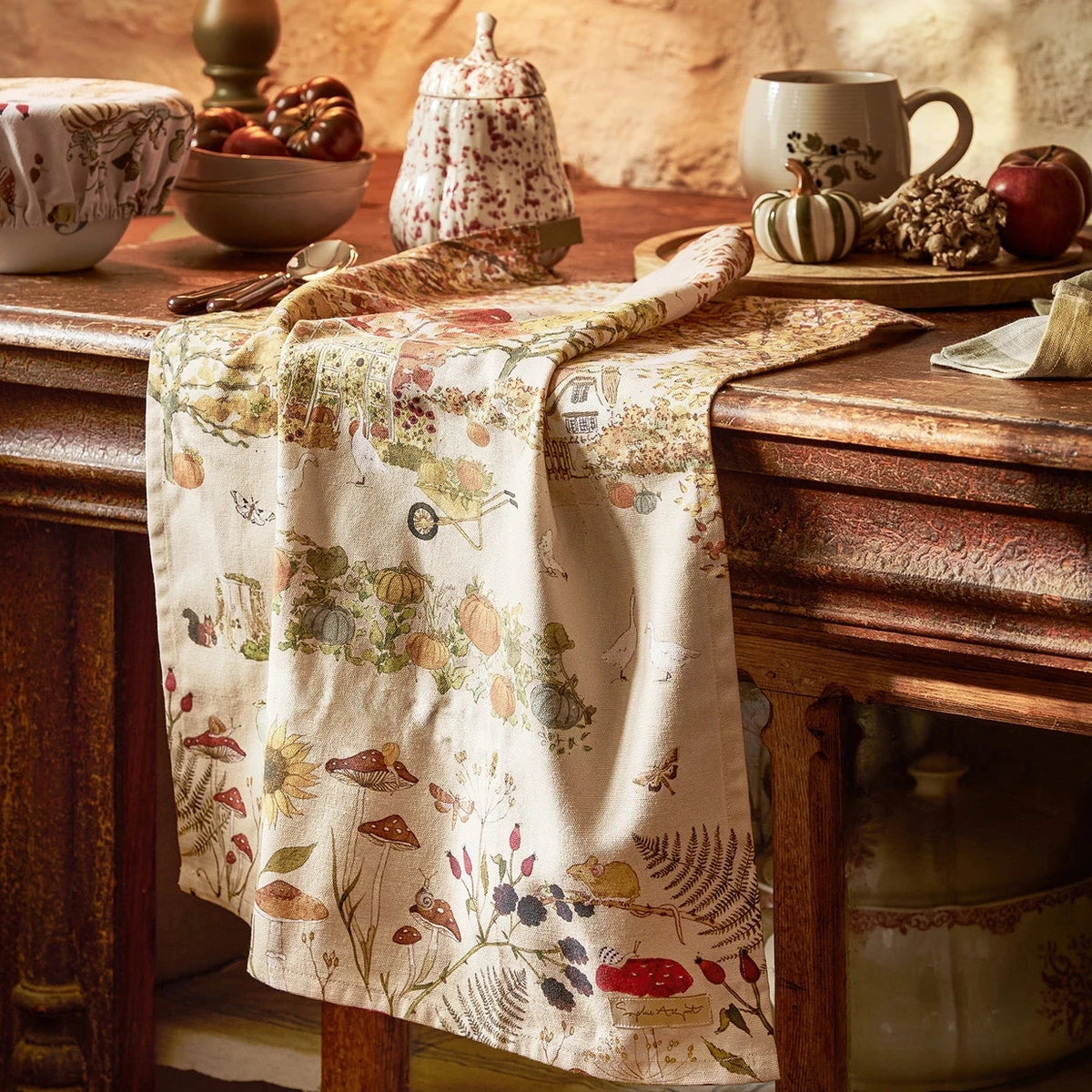 A rustic tea towel with a vintage botanical and mushroom pattern draped over a dark wooden table in a warm, cozy kitchen setting.