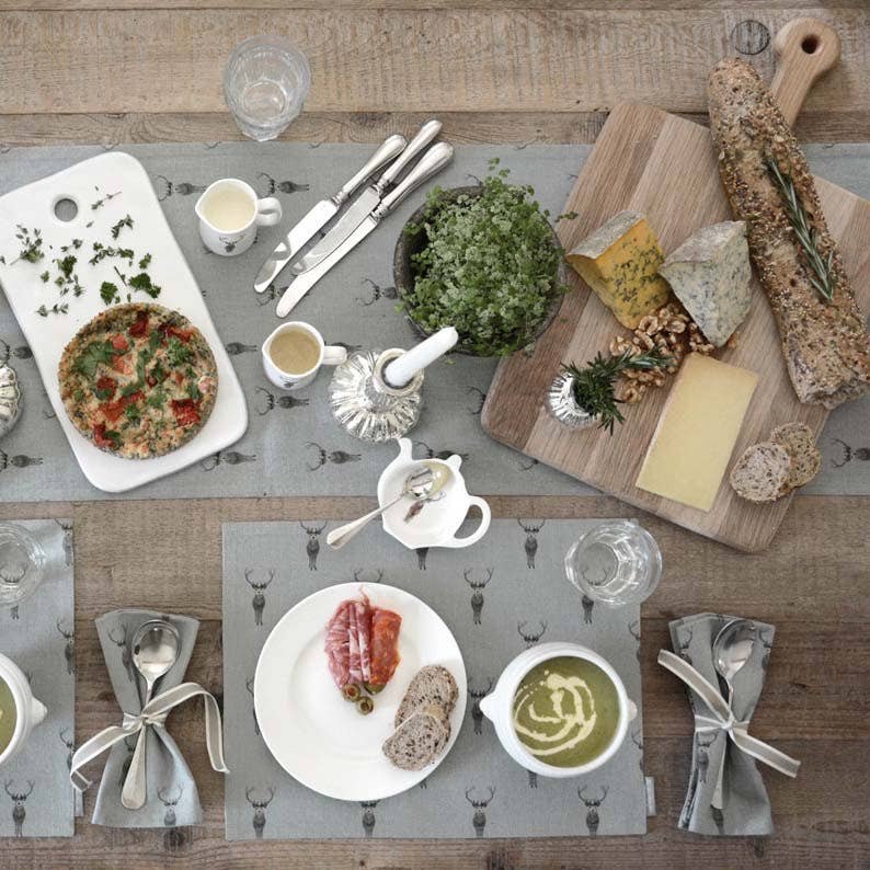 An overhead styled food and tableware scene on a rustic wooden table. Multiple white plates, bowls of soup and greens, cheese, bread, crackers, silverware, cups, and gray placemats with deer motifs are arranged neatly.